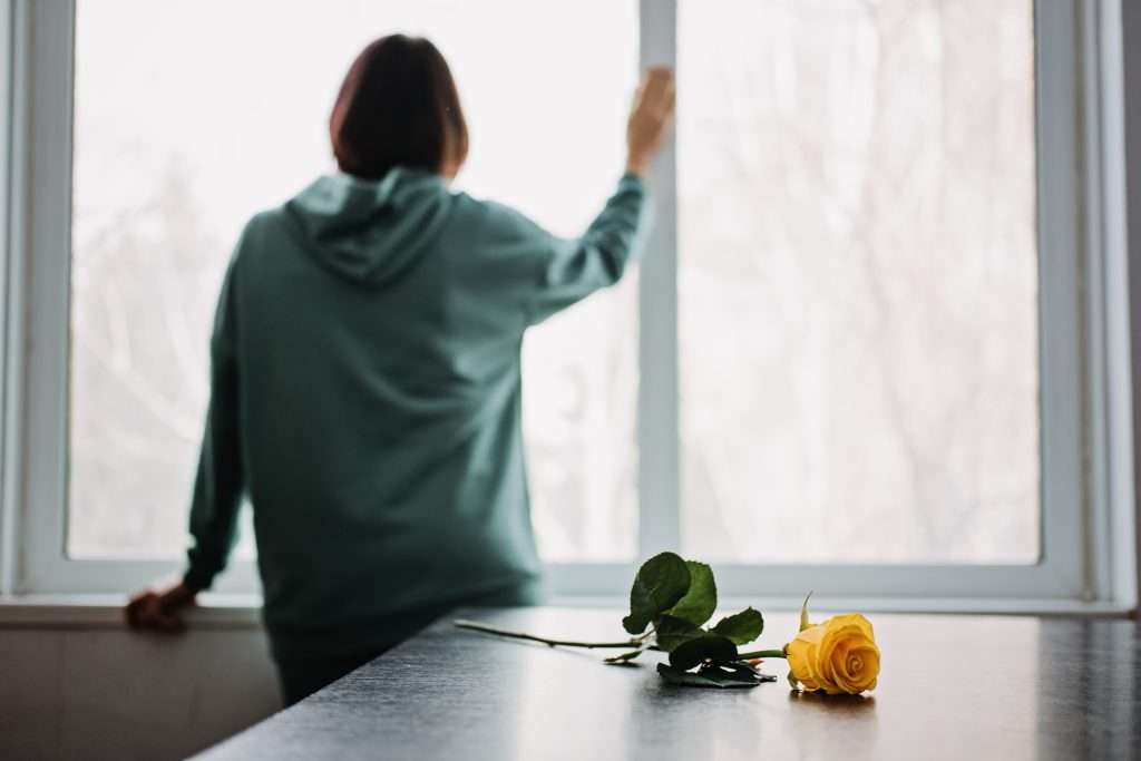 Back view of unhappy sad alone young woman with yellow rose flower at home. Borderline Personality Disorder